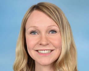 Eine lächelnde Frau mit langen, blonden Haaren trägt eine Halskette. Der Hintergrund ist hellblau.