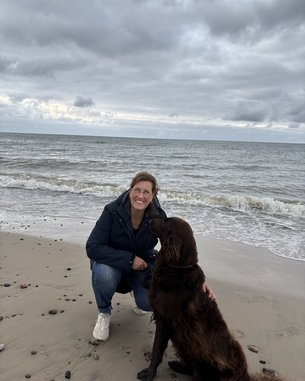 Eine Frau kniet am Strand und lächelt, während sie ihren braunen Hund streichelt. Im Hintergrund sind Wellen und bewölkter Himmel zu sehen.