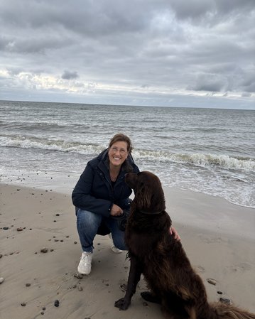 Eine Frau kniet am Strand und lächelt, während sie ihren braunen Hund streichelt. Im Hintergrund sind Wellen und bewölkter Himmel zu sehen.