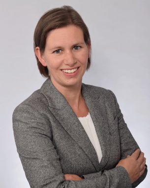 Eine Frau mit kurzen, braunen Haaren lächelt und steht mit verschränkten Armen. Sie trägt einen grauen Blazer über einem weißen Oberteil. Der Hintergrund ist hell und neutral.