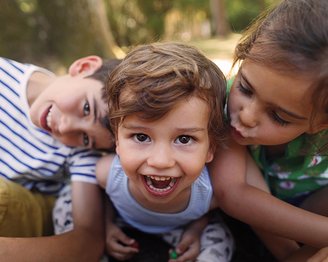 Drei Kinder posieren fröhlich zusammen, wobei ein Junge in der Mitte lächelt und die anderen beiden ihn umarmen. Eines der Kinder gibt ihm einen Kuss auf die Wange.