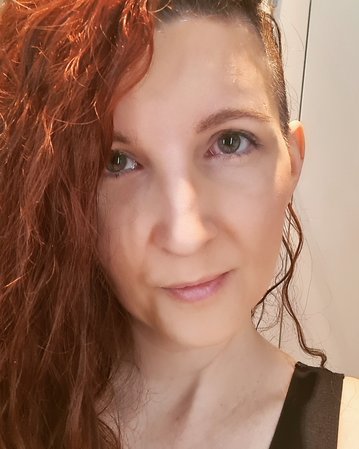 Eine Person mit langen, lockigen, roten Haaren schaut direkt in die Kamera. Sie trägt ein schwarzes Oberteil und hat einen neutralen Gesichtsausdruck.