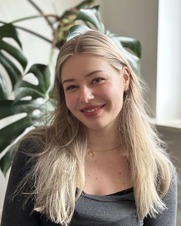 Eine junge Frau mit langen, blonden Haaren lächelt in die Kamera, während sie vor einer Fensterbank mit einer Pflanze sitzt.