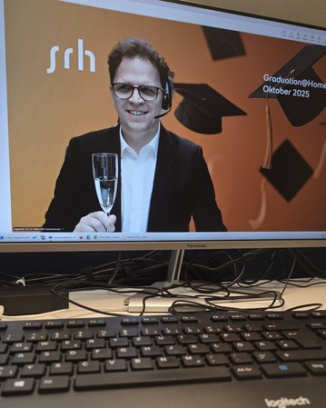 Mann mit Brille und Headset hält ein Glas in der Hand, vor einem Bildschirm mit dem Text "Graduation@Home Oktober 2023".