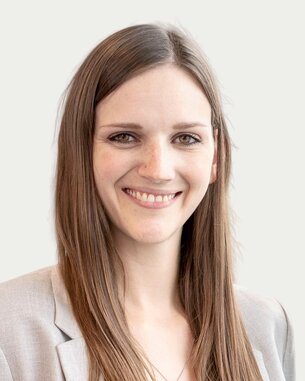 Eine Frau mit langen, glatten Haaren trägt einen hellen Blazer und lächelt freundlich in die Kamera. Der Hintergrund ist neutral.