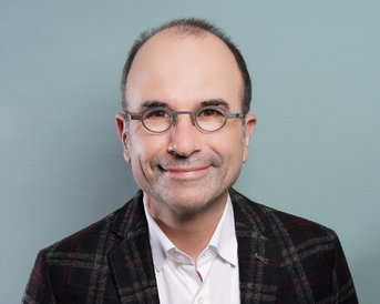 Ein Mann mit Brille und einem karierten Blazer lächelt in die Kamera. Der Hintergrund ist einfarbig und hell.