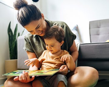 Bindungstheorie Bowlby Eine Mutter liest einem kleinen Kind ein Buch vor, während sie auf dem Boden sitzt. Beide lächeln und zeigen Interesse an der Geschichte.