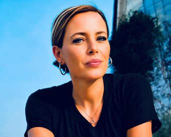 Eine Frau mit kurzen, braunen Haaren und Ohrringen schaut direkt in die Kamera, trägt ein schwarzes T-Shirt und hat einen neutralen Gesichtsausdruck.