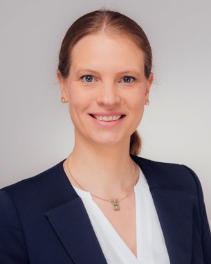 Eine Frau mit langen, blonden Haaren und einem freundlichen Lächeln trägt einen dunkelblauen Blazer über einem weißen Oberteil. Der Hintergrund ist neutral und hell.