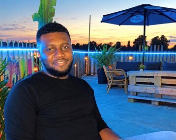 Ein Mann sitzt entspannt auf einer Terrasse, hält ein Smartphone in der Hand, umgeben von Pflanzen und einer gemütlichen Atmosphäre bei Sonnenuntergang.