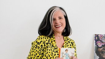 Eine Frau mit langen, grauen Haaren trägt ein gelbes, gepunktetes Hemd und hält ein Buch in der Hand. Neben ihr stehen eine Statue und eine Fotografie einer älteren Frau mit einer Maske.