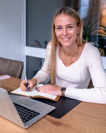 Eine junge Frau sitzt an einem Tisch, schreibt in ein Notizbuch und schaut lächelnd in die Kamera. Ein Laptop und eine Pflanze sind sichtbar.