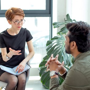 Eine Frau mit kurzen, roten Haaren und Brille sitzt auf einem Stuhl und spricht mit einem Mann, der ihr gegenüber sitzt. Beide scheinen in ein Gespräch vertieft zu sein.