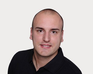 Ein Mann mit kurzem, glattem Haar trägt ein schwarzes Polohemd und schaut direkt in die Kamera. Der Hintergrund ist neutral.