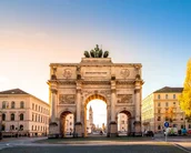 Das Siegestor in München, ein monumentales Triumphbogen mit einer Skulptur von vier Pferden auf dem Dach, umgeben von historischen Gebäuden und einer klaren blauen Himmel.