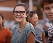Eine junge Frau mit Brille lächelt in die Kamera, umgeben von anderen Jugendlichen. Sie trägt ein graues T-Shirt und hat lange, lockige Haare.