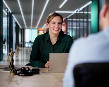 Eine Frau in einem grünen Hemd sitzt an einem Tisch und spricht mit einem Mann. Auf dem Tisch steht eine Statue der Justitia.