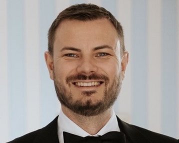 Ein Mann mit Bart trägt einen schwarzen Anzug und eine Fliege. Er lächelt freundlich in die Kamera, vor einem Hintergrund mit blauen Streifen.