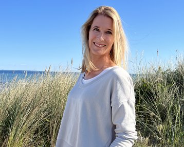 Eine Frau mit langen, blonden Haaren steht an einem Strand, umgeben von Gras und einem weißen Zaun. Sie trägt einen grauen Pullover und Jeans, während sie in die Kamera lächelt.