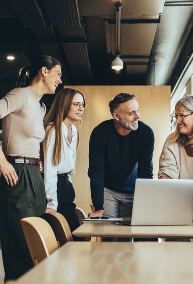 Sechs Personen stehen um einen Tisch in einem modernen Büro und schauen auf einen Laptop. Sie scheinen eine Diskussion oder Präsentation zu führen.