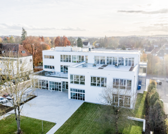 Moderne, mehrstöckige Gebäude mit großen Fenstern und einem flachen Dach, umgeben von Rasenflächen und Bäumen. Die Architektur ist schlicht und funktional, mit einem klaren, weißen Außenanstrich.