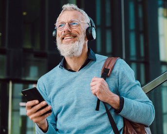 Ein älterer Mann mit Brille und Kopfhörern lächelt, während er ein Smartphone in der Hand hält. Er trägt einen hellblauen Pullover und eine braune Tasche.
