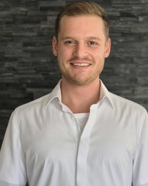 Mann mit kurzem, blondem Haar trägt ein weißes Hemd und lächelt vor einer dunklen Steinwand.