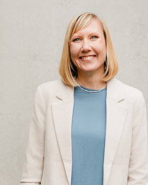 Eine lächelnde Frau mit blonden Haaren trägt einen hellen Blazer und ein blaues Oberteil. Sie steht vor einer grauen Wand mit Pflanzen im Vordergrund.