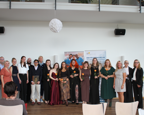 Gruppe von Menschen in festlicher Kleidung, die auf einer Veranstaltung zusammenstehen, einige halten Blumen. Im Hintergrund ein Banner mit dem Text „DER PFLEGEMANAGER“.