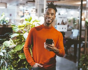Ein Mann mit einem breiten Lächeln trägt einen orangefarbenen Pullover und hält ein Smartphone in der Hand, umgeben von grünen Pflanzen in einem modernen Innenraum.