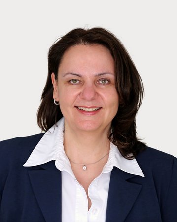 Eine Frau mit langen, dunklen Haaren trägt ein dunkelblaues Blazer und ein weißes Hemd. Sie lächelt freundlich in die Kamera. Der Hintergrund ist neutral und hell.
