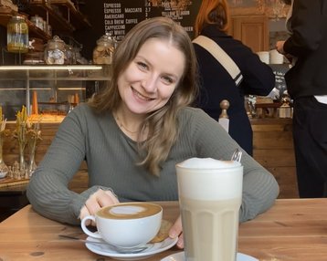 Eine lächelnde Frau sitzt an einem Tisch in einem Café, vor ihr stehen eine Tasse Kaffee und ein Glas Latte Macchiato, begleitet von einem Keks. Im Hintergrund sind weitere Café-Möbel und Gäste sichtbar.