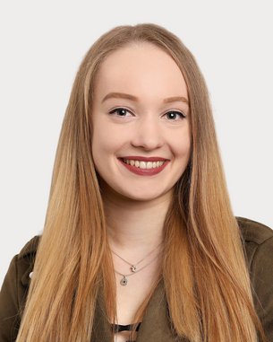 Eine junge Frau mit langen, blonden Haaren und einem freundlichen Lächeln trägt eine grüne Jacke. Der Hintergrund ist neutral und hell.
