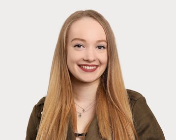 Eine junge Frau mit langen, blonden Haaren und einem freundlichen Lächeln trägt eine grüne Jacke. Der Hintergrund ist neutral und hell.