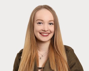 Eine junge Frau mit langen, blonden Haaren und einem freundlichen Lächeln trägt eine grüne Jacke. Der Hintergrund ist neutral und hell.