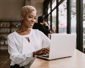 Eine Frau mit kurzen, lockigen Haaren sitzt an einem Tisch und arbeitet an einem Laptop. Sie trägt eine weiße Bluse und lächelt, während sie auf den Bildschirm schaut. Im Hintergrund ist eine andere Person zu sehen.