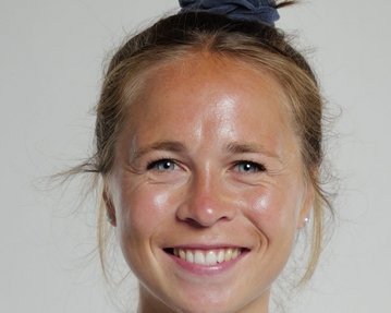 Porträt einer sportlichen Frau mit einem Dutt, die ein schwarzes Trikot mit einem Vereinslogo trägt. Sie lächelt freundlich in die Kamera.
