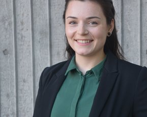 Eine junge Frau mit dunklen Haaren trägt ein grünes Hemd und einen schwarzen Blazer. Sie lächelt und steht vor einer strukturierten Wand.