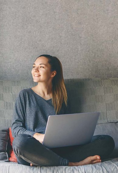 Eine Frau sitzt auf einem Sofa, lächelt und arbeitet an einem Laptop. Sie trägt einen grauen Pullover und hat ihre Beine angezogen. Im Hintergrund ist eine Stehlampe und ein gemusterter Vorhang zu sehen.