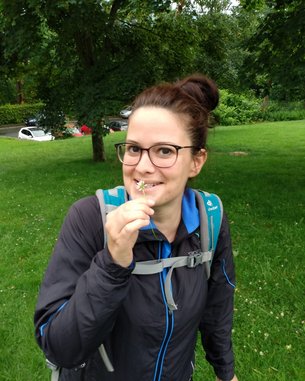 Eine Person mit Brille hält eine kleine Blume in der Hand und lächelt. Sie trägt eine Jacke und einen Rucksack und steht auf einer grünen Wiese.