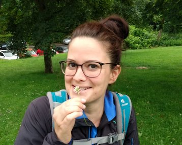 Eine Person mit Brille hält eine kleine Blume in der Hand und lächelt. Sie trägt eine Jacke und einen Rucksack und steht auf einer grünen Wiese.