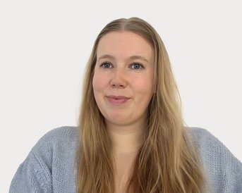Eine Frau mit langen, blonden Haaren trägt einen grauen Pullover und schaut in die Kamera. Der Hintergrund ist neutral und hell.