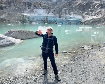 Eine Person steht am Ufer eines Gletschersees, trägt eine schwarze Jacke und eine Mütze, und winkt in die Kamera. Im Hintergrund sind Gletscher und Eisberge sichtbar.