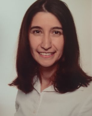 Eine Frau mit langen, dunklen Haaren und einem freundlichen Lächeln trägt ein helles Hemd. Der Hintergrund ist neutral und hell.