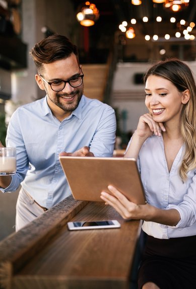 Drei Personen in einem modernen Café betrachten gemeinsam ein Tablet. Zwei Männer, einer mit Brille, halten eine Tasse, während die Frau lächelt und auf den Bildschirm zeigt.