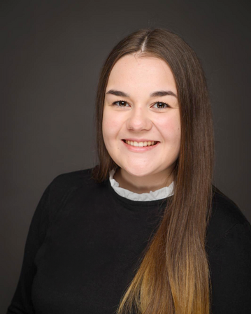 Eine junge Frau mit langen, glatten Haaren und einem freundlichen Lächeln trägt ein schwarzes Oberteil und einen weißen Kragen.