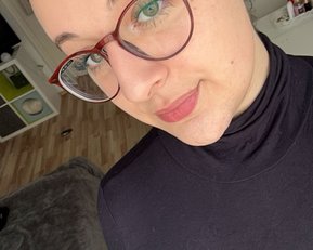 Eine Person mit kurzen, glatten Haaren und Brille trägt ein schwarzes Oberteil. Sie steht in einem hellen Raum mit Möbeln im Hintergrund.