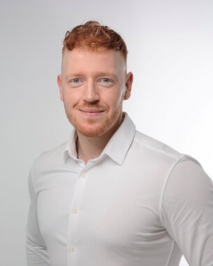 Ein Mann mit rotem Haar und Bart trägt ein weißes Hemd und lächelt in die Kamera. Der Hintergrund ist hell und neutral.