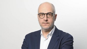 Ein Mann mit Brille und kurzem, grauem Bart trägt einen dunkelblauen Anzug und ein weißes Hemd. Er steht mit verschränkten Armen vor einem hellen Hintergrund.