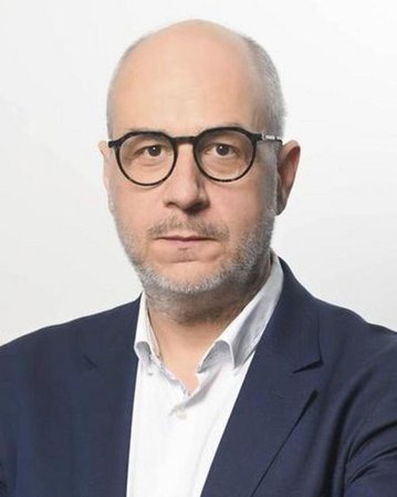 Ein Mann mit Brille und kurzem, grauem Bart trägt einen dunkelblauen Anzug und ein weißes Hemd. Er steht mit verschränkten Armen vor einem hellen Hintergrund.